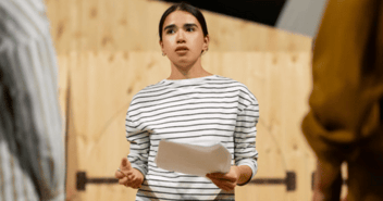 Una mujer joven sostiene un documento mientras habla con determinación frente a un grupo, destacando en un ambiente profesional y enfocado en la comunicación.