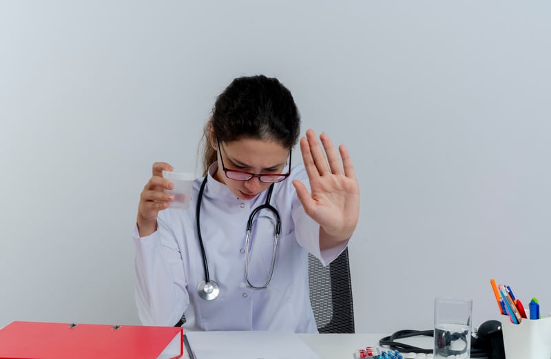 joven-doctora-disgustada-con-bata-medica-y-estetoscopio-y-gafas-sentado-en-el-escritorio-con-herramientas-medicas-sosteniendo-vaso-medico-mirando-hacia-abajo-haciendo-gesto-de-parada-aislado